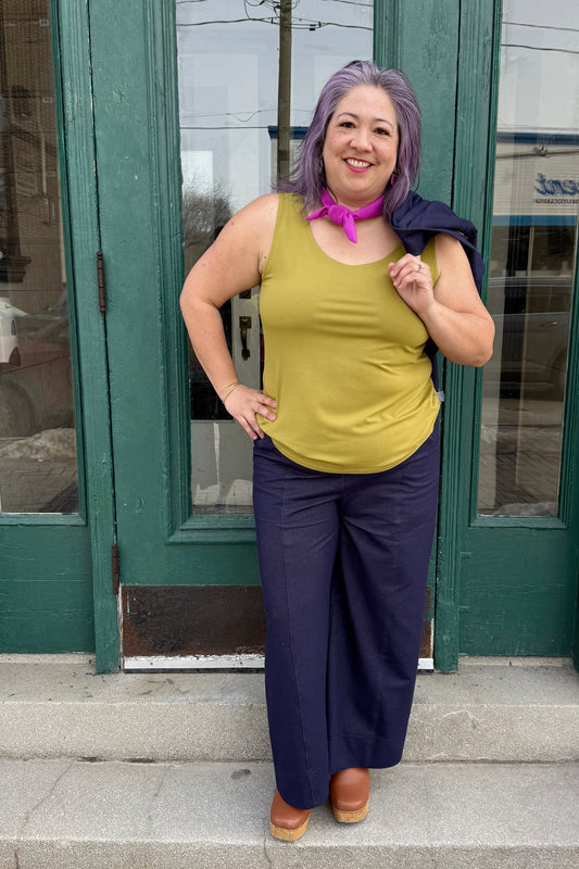 A woman wearing the Brooke Knit Trousers by Julei in Navy Knit, wide-leg pants with a pull-on waist, front slant pockets, and back patch pockets. She is wearing them a green tank and holding a navy jacket over her shoulder. She is standing outside in front of a green door. 