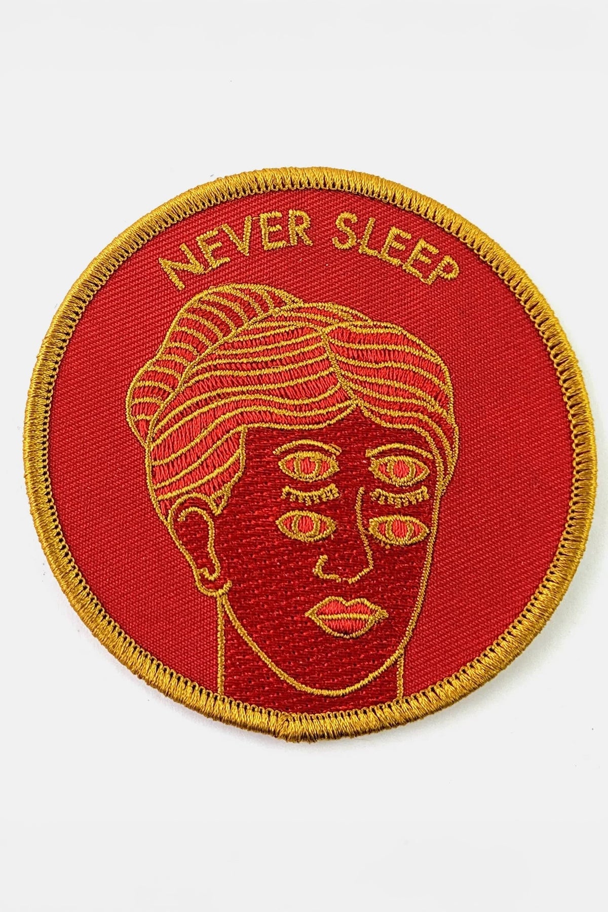 The Never Sleep Patch from Badaboöm Studio, circular embroidered patch with a red background, gold border, and a stylized face with two sets of open eyes. The words 'NEVER SLEEP' are embroidered in gold at the top.