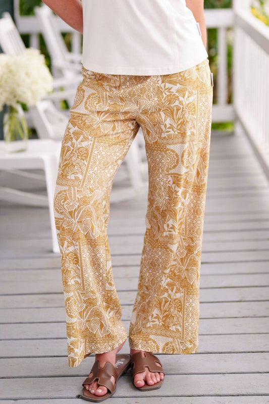 Waist down view of a woman wearing the Mango Pants from Rien ne se Perd in Ocre/Ivory tropical print, palazzo pants with an elastic waist and pockets. She is wearing them with a white top and standing on a porch. 