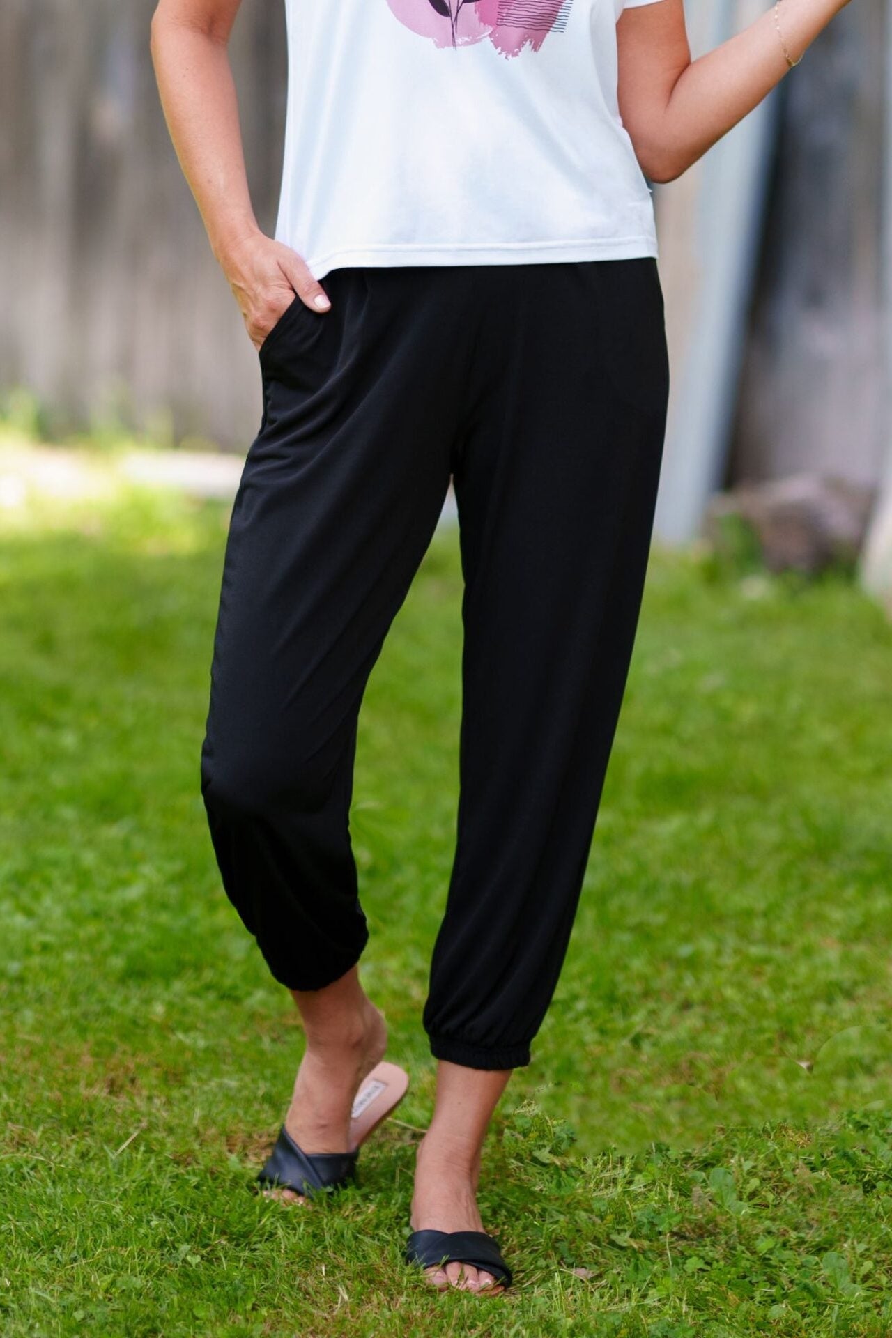 Waist down view of a woman wearing the Pina 7/8 Pants by Rien ne se Perd in Bloack, with a pull on waist, front pleats, cuffed legs, and a pockets. She is wearing them with a white top and standing on the grass.