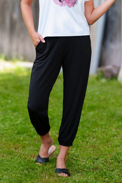 Waist down view of a woman wearing the Pina 7/8 Pants by Rien ne se Perd in Bloack, with a pull on waist, front pleats, cuffed legs, and a pockets. She is wearing them with a white top and standing on the grass.