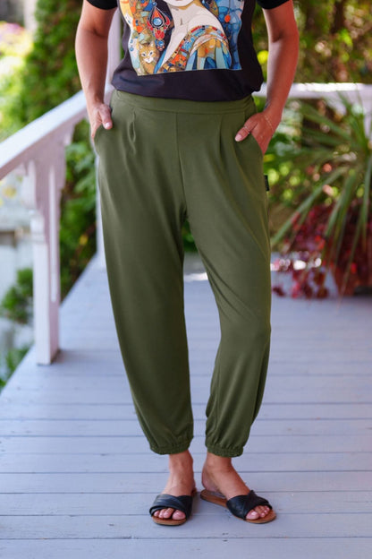 Waist down view of a woman wearing the Pina 7/8 Pants by Rien ne se Perd in Olive, with a pull on waist, front pleats, cuffed legs, and a pockets. She is wearing them with a printed top and standing on a porch with greenery in the background. 