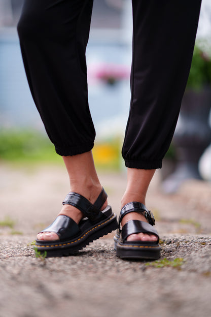 Waist down view of a woman wearing the Pina 7/8 Pants by Rien ne se Perd in Black, with a pull on waist, front pleats, cuffed legs, and a pockets. She is standing outside. 