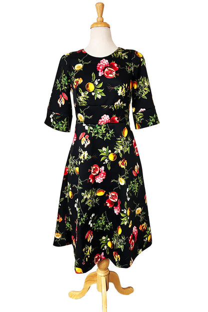 The Pomegranate and Friends Dress by Desserts and Skirts, a black dress with a fruit pattern with half sleeves, a fit and flare shape, and a below the knee length, is shown on a mannequin against a white background 