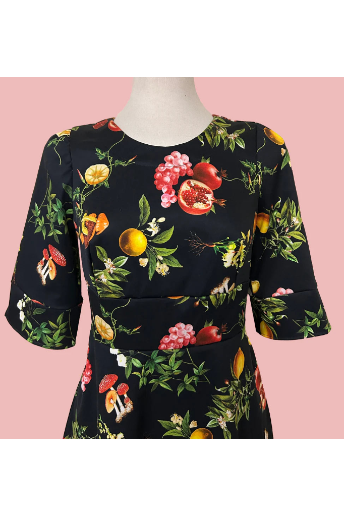  A close up view of the Pomegranate and Friends Dress by Desserts and Skirts, a black dress with a fruit pattern with half sleeves, a fit and flare shape, and a below the knee length, is shown on a mannequin against a pink background.
