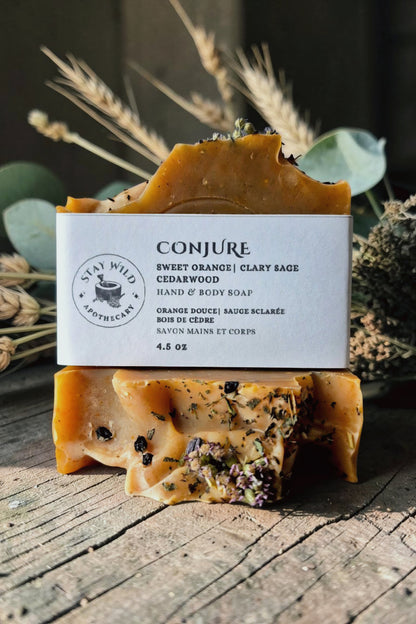 Two stacked bars of hand and body soap with a white paper label reading 'CONJURE' and 'Sweet Orange | Clary Sage Cedarwood', photographed on a rustic wooden surface with dried wheat and greenery in the background.