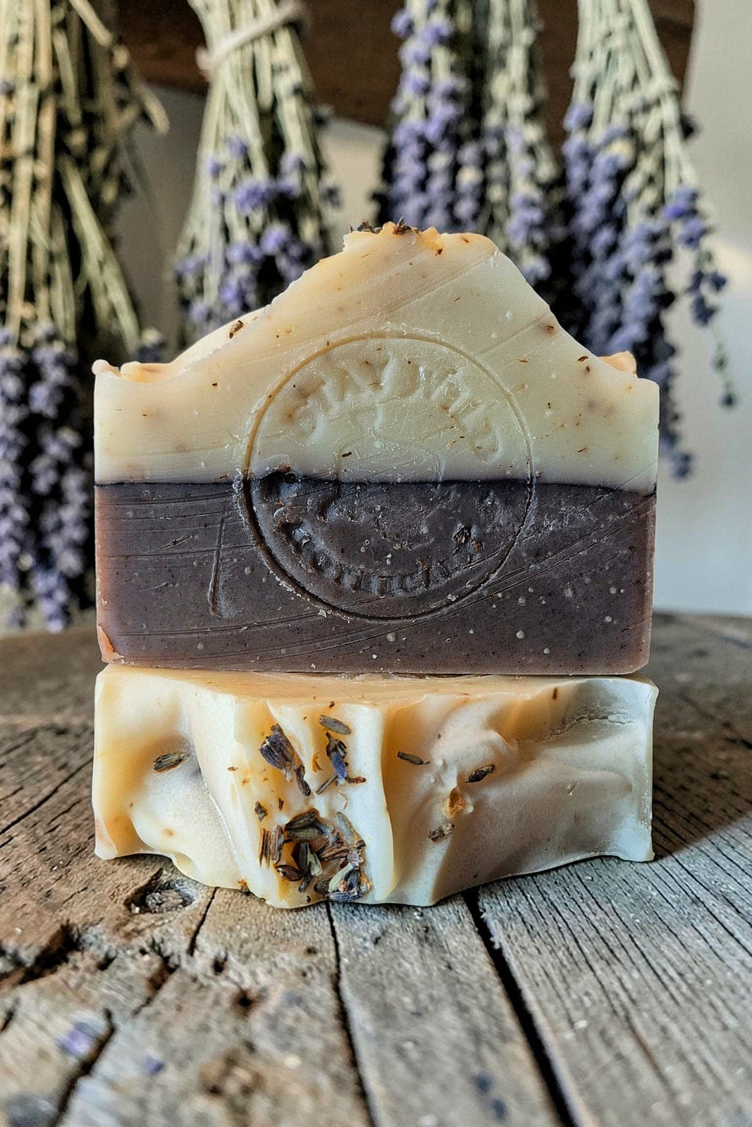 Two stacked bars of Witch's Garden soap bar from Stay Wild Apothecary, with a cream-coloured top half and a dark brown bottom half, displayed on a wooden table with dried lavender hanging behind them. 