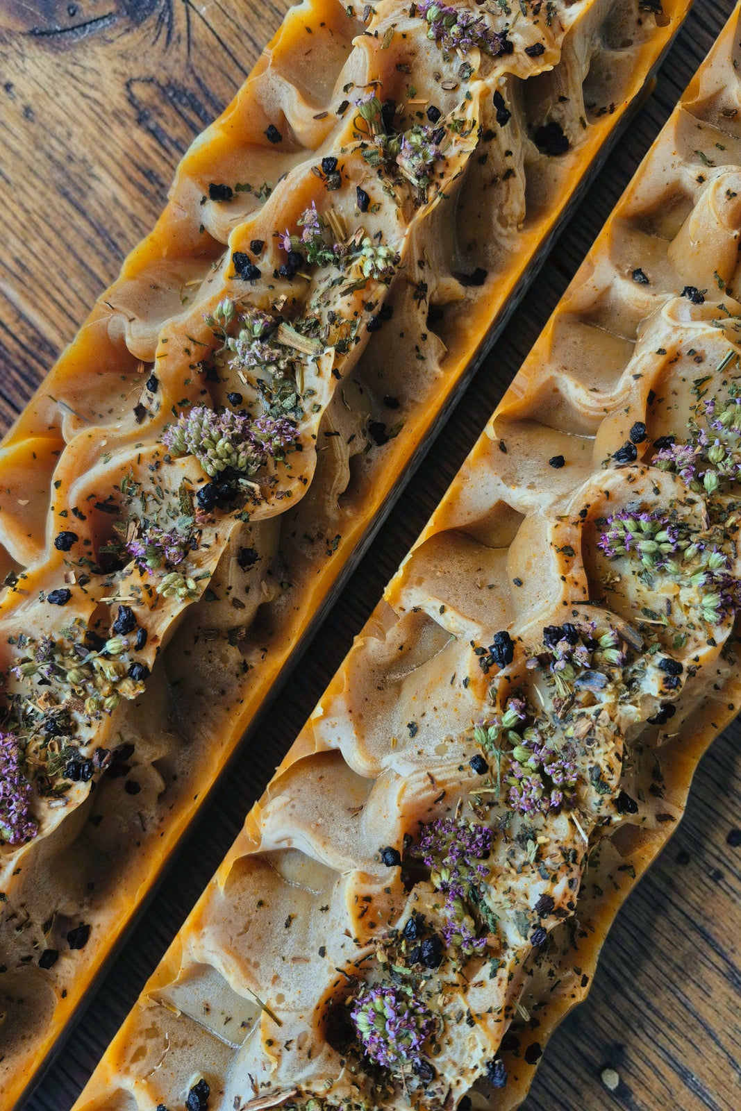 Two long, rectangular bars of artisanal Stay Wild Apothecary Conjure soap in a diagonal overhead view on a rustic wooden surface. The soap has a wavy, layered texture in warm tan and brown tones, topped with dried herbs, seeds, and small purple flowers