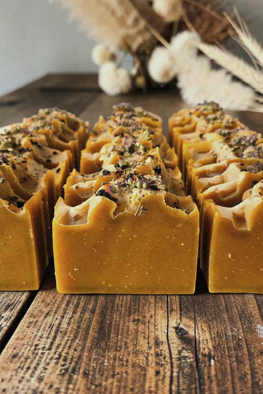 Several rectangular bars of handmade Stay Wild Apothecary Conjure soap, golden-yellow soap with textured tops and dried botanical toppings, arranged on a rustic wooden surface with dried grasses in the background.