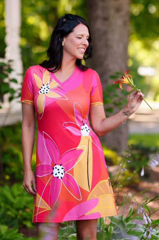 A woman wearing the Aperol Dress from Rien ne se Perd, a short flared dress with a V-neck, short sleeves, and a colourful Pink floral pattern She is holding a flower in a garden setting.