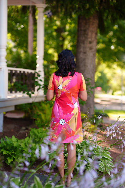 Back view of a woman wearing the Aperol Dress from Rien ne se Perd, a short flared dress with a V-neck, short sleeves, and a colourful Pink floral pattern She is standing in a garden setting.