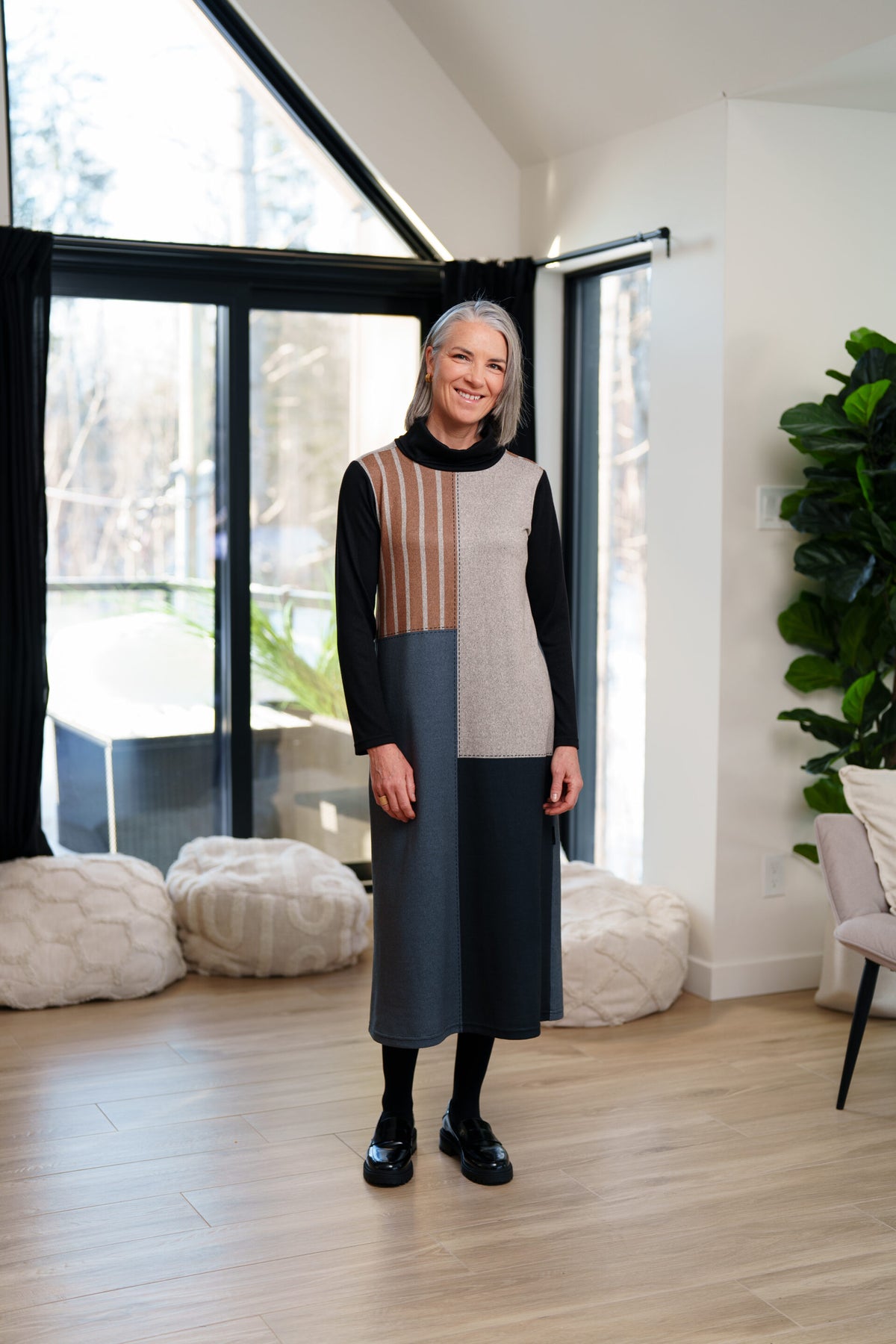 A woman wearing the Bonaventure Dress by Rien ne se Perd, a colour-blocked turtleneck sweater dress with panels of solid grey, blue and black, and brown stripes. She is standing in a room with bean bag chairs behind her. 
