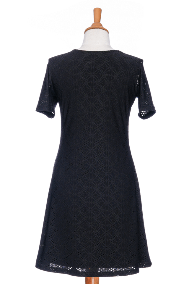 A back view of the Litchi Dress by Rien en se Perd in Black, a semi-fitted short dress with short sleeves and an openwork daisy fabric, is shown on a mannequin against a white background. 