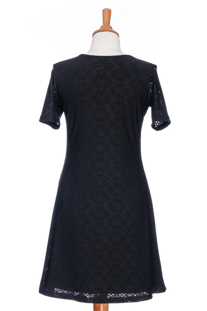 A back view of the Litchi Dress by Rien en se Perd in Black, a semi-fitted short dress with short sleeves and an openwork daisy fabric, is shown on a mannequin against a white background. 