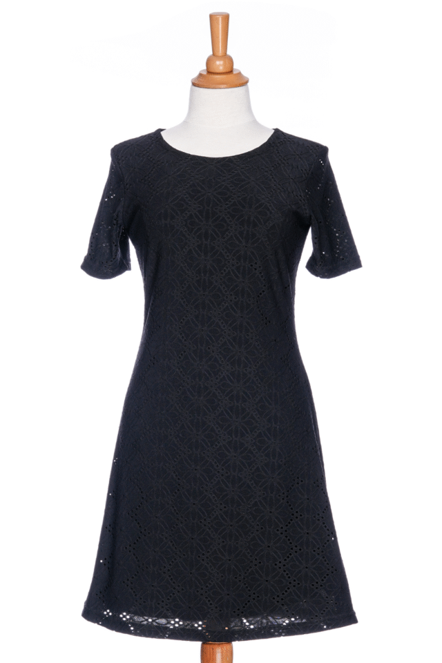 The Litchi Dress by Rien en se Perd in Black, a semi-fitted short dress with short sleeves and an openwork daisy fabric, is shown on a mannequin against a white background. 