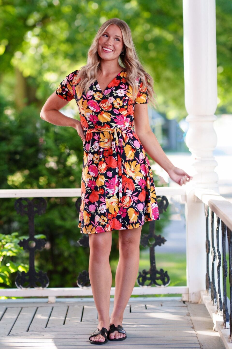A woman wearing the Margarita Dress by Rien ne se Perd in Red/Orange floral print, with a faux wrap neckline, short sleeves, and short a-line skirt. She is standing on a porch. 