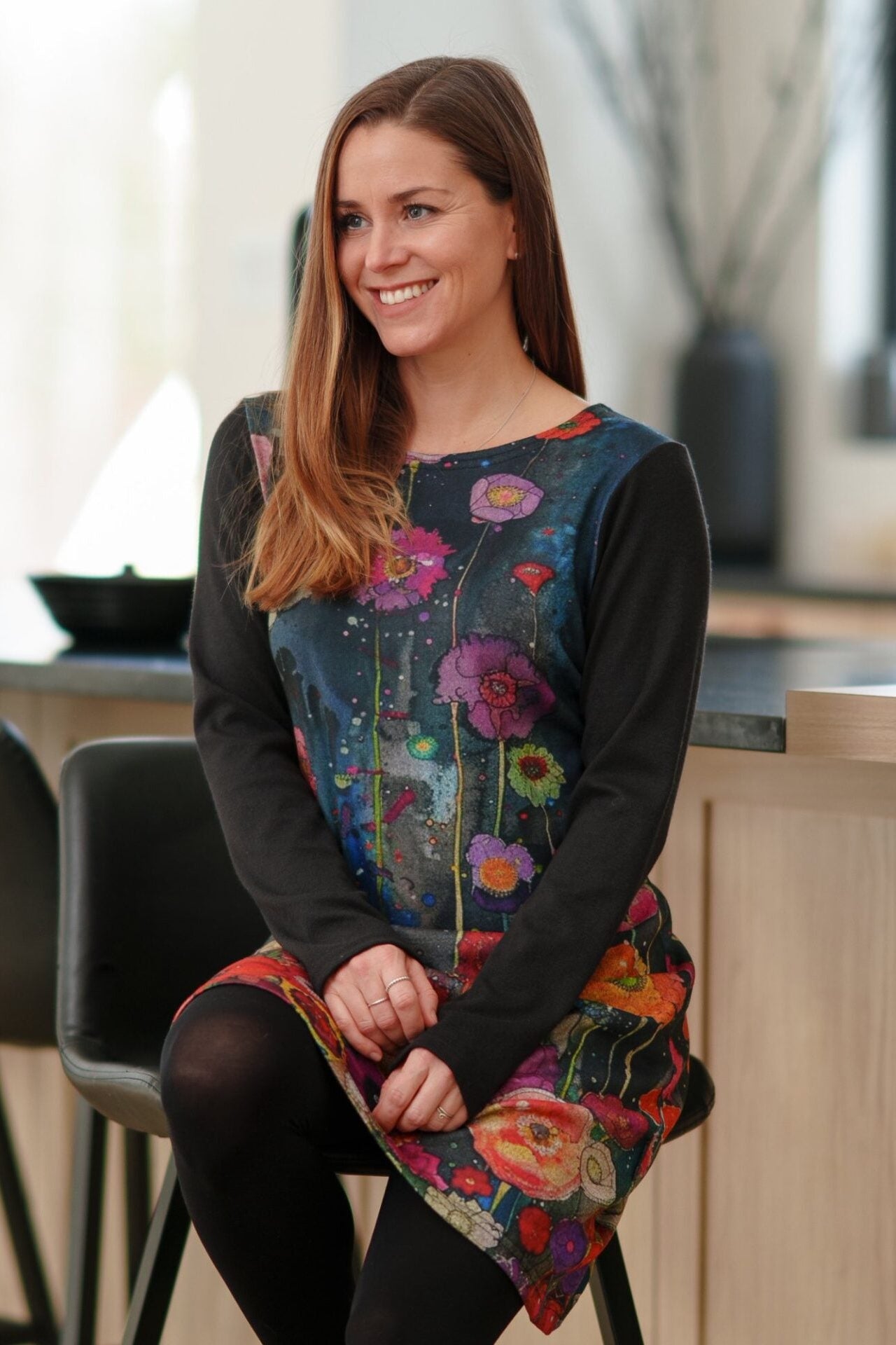 A woman wearing the Mistassini Dress by Rien ne se Perd in Floral Watercolour, an A-line knee-length dress with a vibrant floral print on the main body and contrasting black sleeves. She is sitting on a high stool in a kitchen setting. 