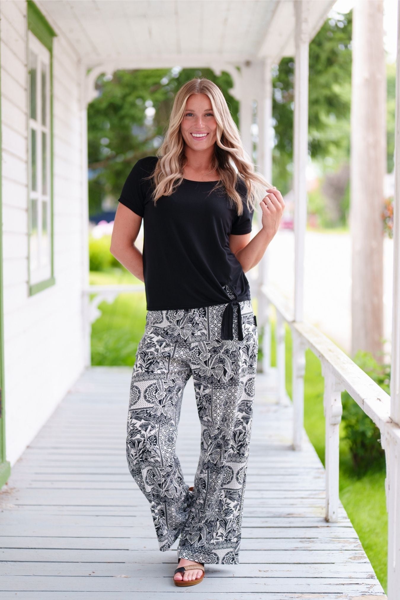 A woman wearing the Mango Pants from Rien ne se Perd in Black/Ivory tropical print, palazzo pants with an elastic waist and pockets. She is wearing them with the Tropical top in Black and standing on a porch. 