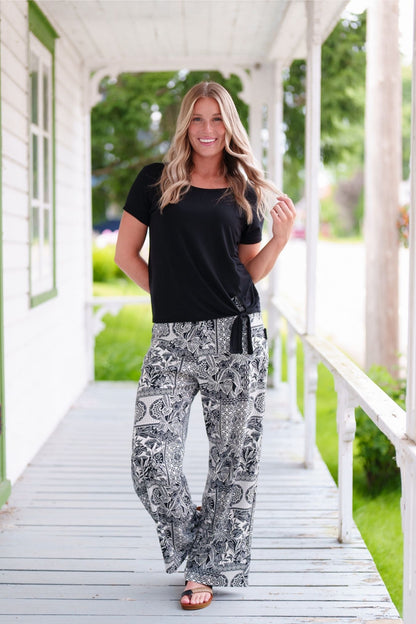 A woman wearing the Mango Pants from Rien ne se Perd in Black/Ivory tropical print, palazzo pants with an elastic waist and pockets. She is wearing them with the Tropical top in Black and standing on a porch. 