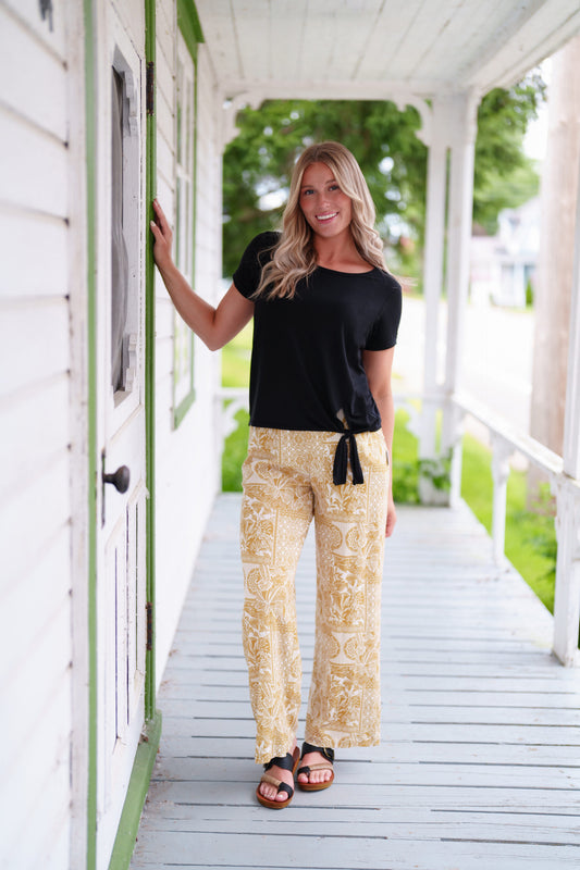 A woman wearing the Mango Pants from Rien ne se Perd in Ocre/Ivory tropical print, palazzo pants with an elastic waist and pockets. She is wearing them with the Tropical top in Black and standing on a porch. 