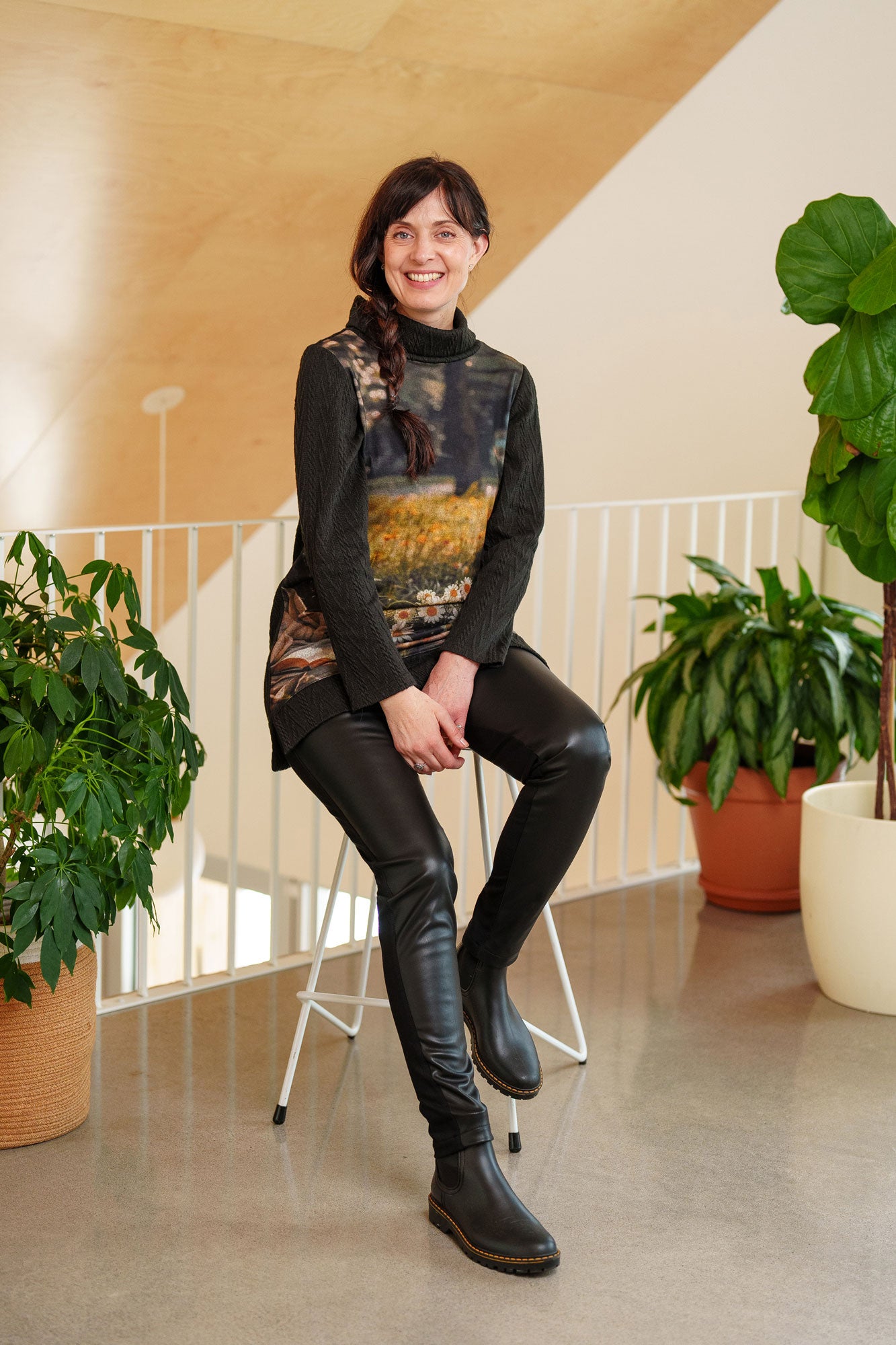 A woman wearing the Resinosa Pants by Rien ne se Perd in Black, slim fitting pants with faux leather at the front and plain fabric at the back, with a wide pull on waistband. She is wearing them with a tunic and sitting on a stool.