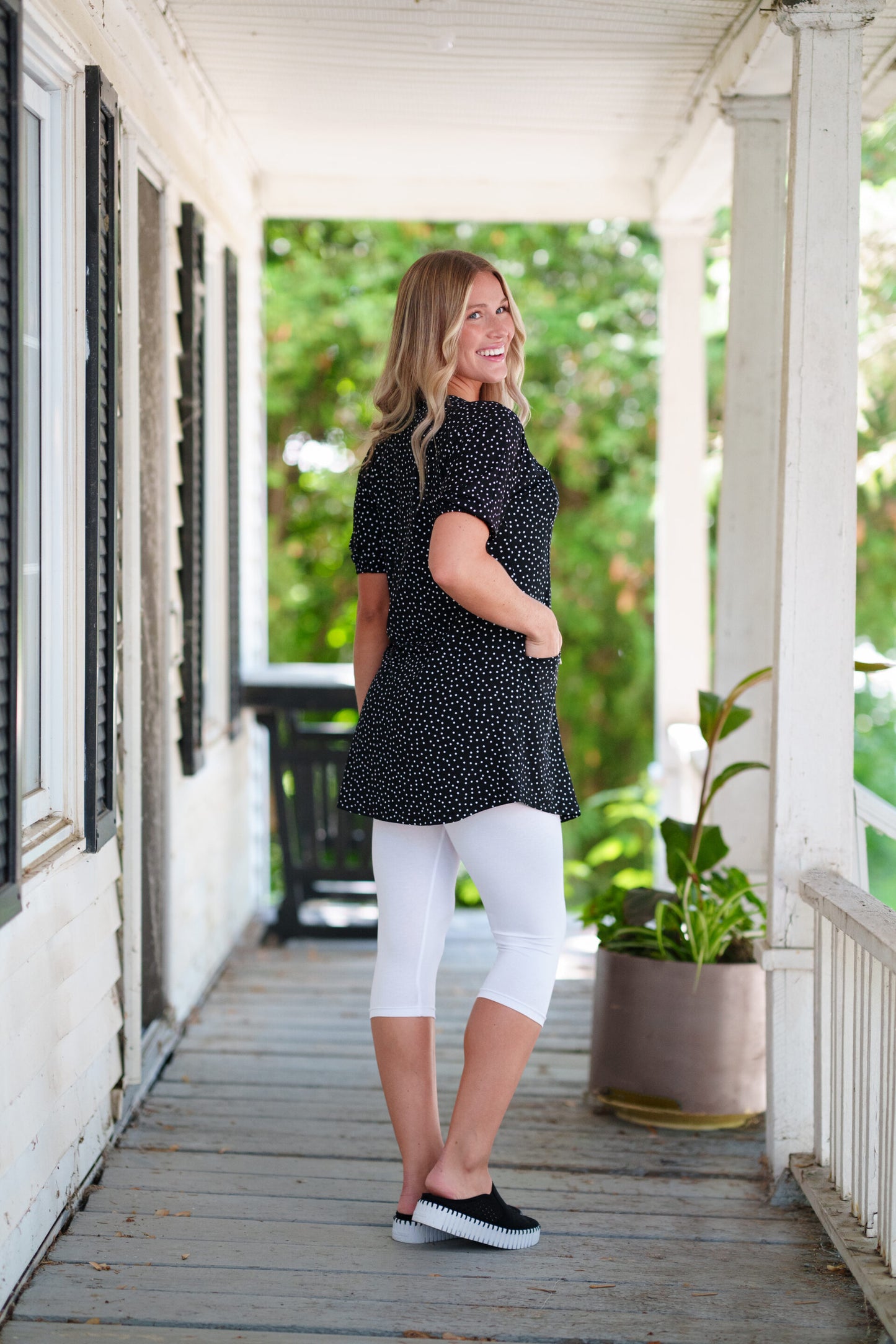 Side view of a woman wearing the Martini Tunic from Rien ne se Perd, a black polka dot piece with full elbow length sleeves with gathered cuffs, and two patch pockets. She is wearing it with white 3/4 leggings and standing outdoors with greenery in the background. 