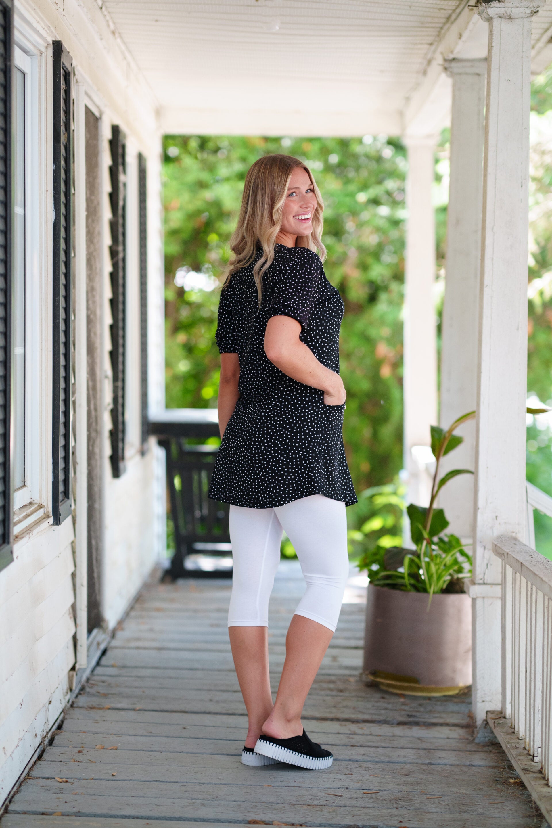 Side view of a woman wearing the Martini Tunic from Rien ne se Perd, a black polka dot piece with full elbow length sleeves with gathered cuffs, and two patch pockets. She is wearing it with white 3/4 leggings and standing outdoors with greenery in the background. 