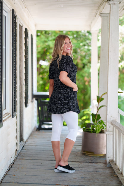 Side view of a woman wearing the Martini Tunic from Rien ne se Perd, a black polka dot piece with full elbow length sleeves with gathered cuffs, and two patch pockets. She is wearing it with white 3/4 leggings and standing outdoors with greenery in the background. 