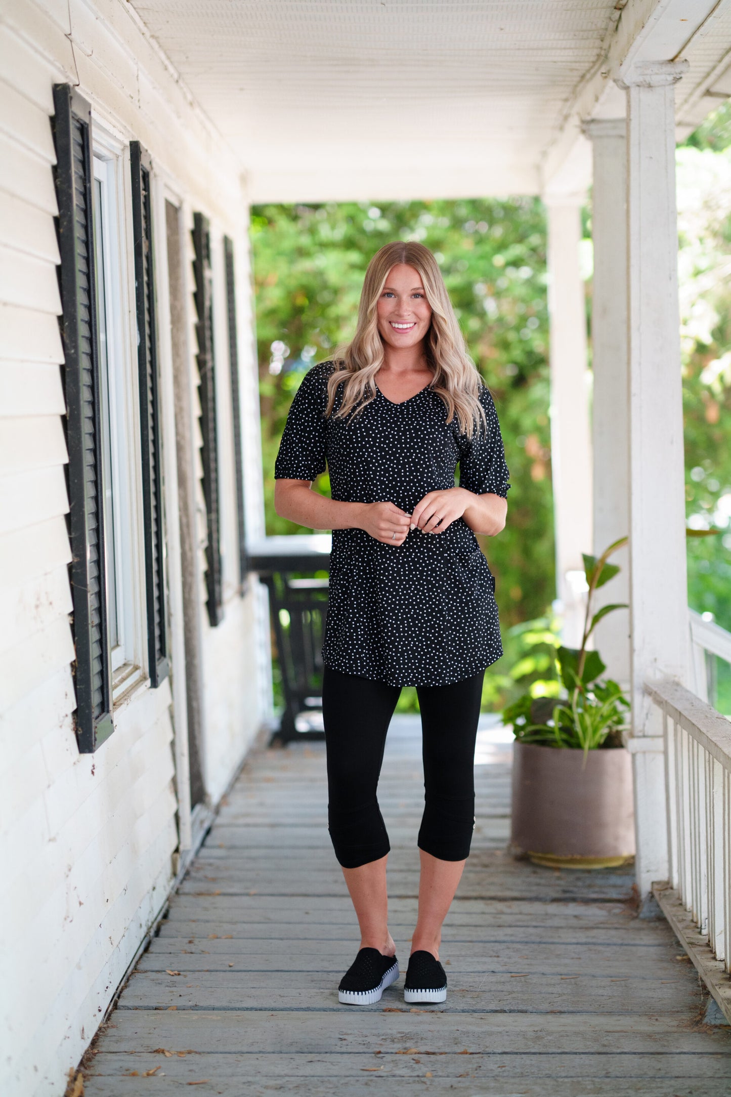 A woman wearing the Martini Tunic from Rien ne se Perd, a black polka dot piece with full elbow length sleeves with gathered cuffs, and two patch pockets. She is wearing it with black 3/4 leggings and standing outdoors on a porch with greenery in the background. 