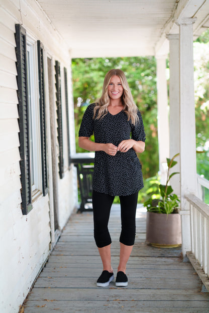A woman wearing the Martini Tunic from Rien ne se Perd, a black polka dot piece with full elbow length sleeves with gathered cuffs, and two patch pockets. She is wearing it with black 3/4 leggings and standing outdoors on a porch with greenery in the background. 