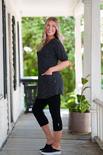 Side view of a woman wearing the Martini Tunic from Rien ne se Perd, a black polka dot piece with full elbow length sleeves with gathered cuffs, and two patch pockets. She is wearing it with black 3/4 leggings and standing outdoors with greenery in the background. 