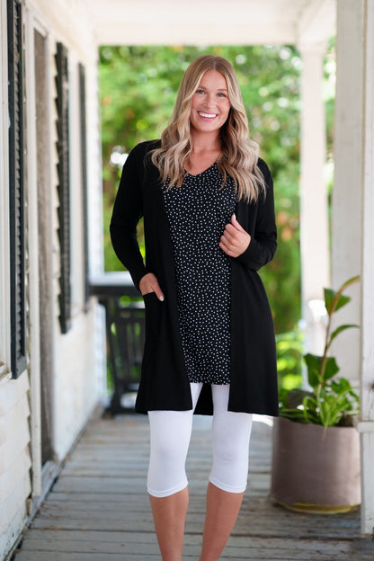 A woman wearing the Martini Tunic from Rien ne se Perd, a black polka dot piece with full elbow length sleeves with gathered cuffs, and two patch pockets. She is wearing it with white 3/4 leggings and a black cardigan and standing outdoors with greenery in the background. 