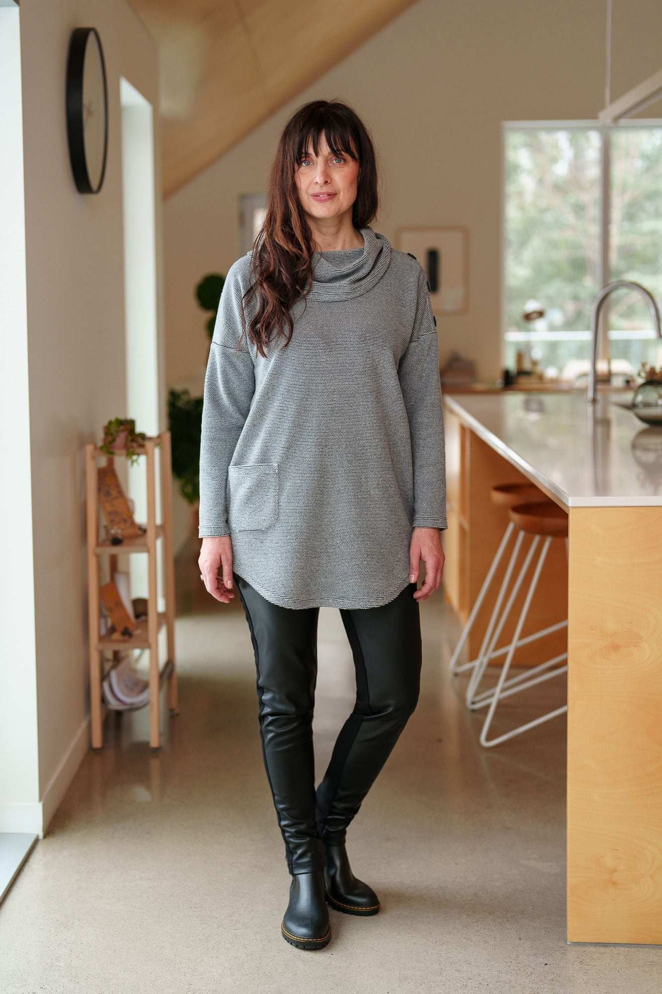 A woman wearing the Resinosa Pants by Rien ne se Perd in Black, slim fitting pants with faux leather at the front and plain fabric at the back, with a wide pull on waistband. She is wearing them with the Myrique Tunic in Grey and standing in a kitchen.