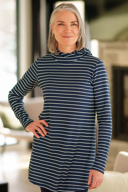 A woman wearing the Rupert Tunic by Rien ne se Perd in Navy/Ivory stripe, with  a crossover neckline and hood, long sleeves, and rounded hem. She is wearing it with black leggings and standing in a living room. 