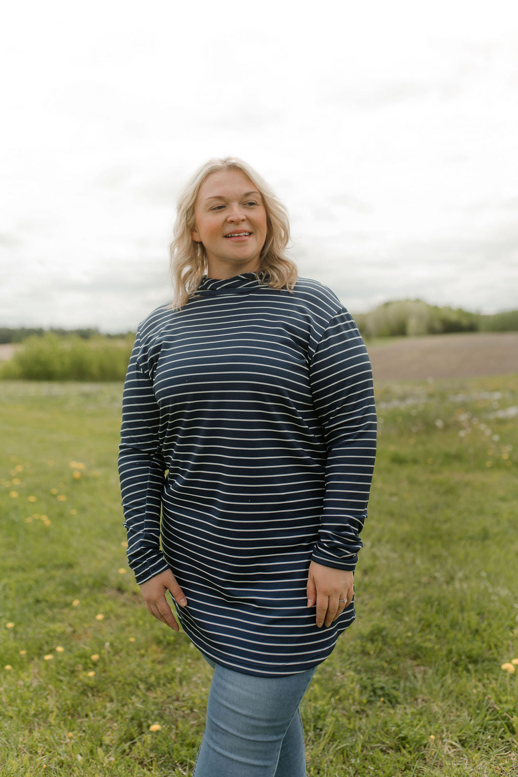 A woman wearing the Rupert Tunic by Rien ne se Perd in Navy/Ivory stripe, with  a crossover neckline and hood, long sleeves, and rounded hem. She is wearing it with jeans and standing outdoors in a field.