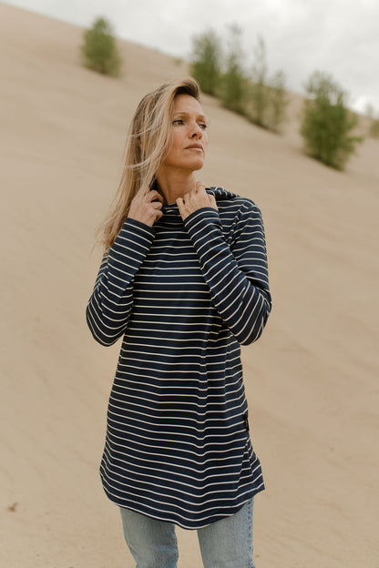 A woman wearing the Rupert Tunic by Rien ne se Perd in Navy/Ivory stripe, with  a crossover neckline and hood, long sleeves, and rounded hem. She is wearing it with jeans and standing outdoors on a sandy hill. 