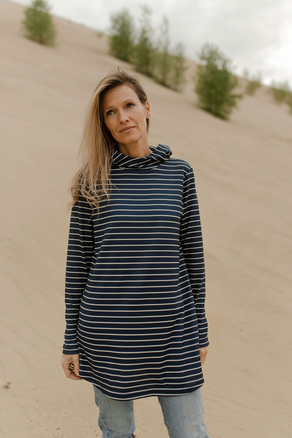 A woman wearing the Rupert Tunic by Rien ne se Perd in Navy/Ivory stripe, with  a crossover neckline and hood, long sleeves, and rounded hem. She is wearing it with jeans and standing outdoors on a sandy hill. 