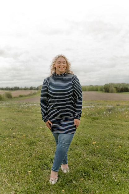 A woman wearing the Rupert Tunic by Rien ne se Perd in Navy/Ivory stripe, with  a crossover neckline and hood, long sleeves, and rounded hem. She is wearing it with jeans and standing outdoors in a field.