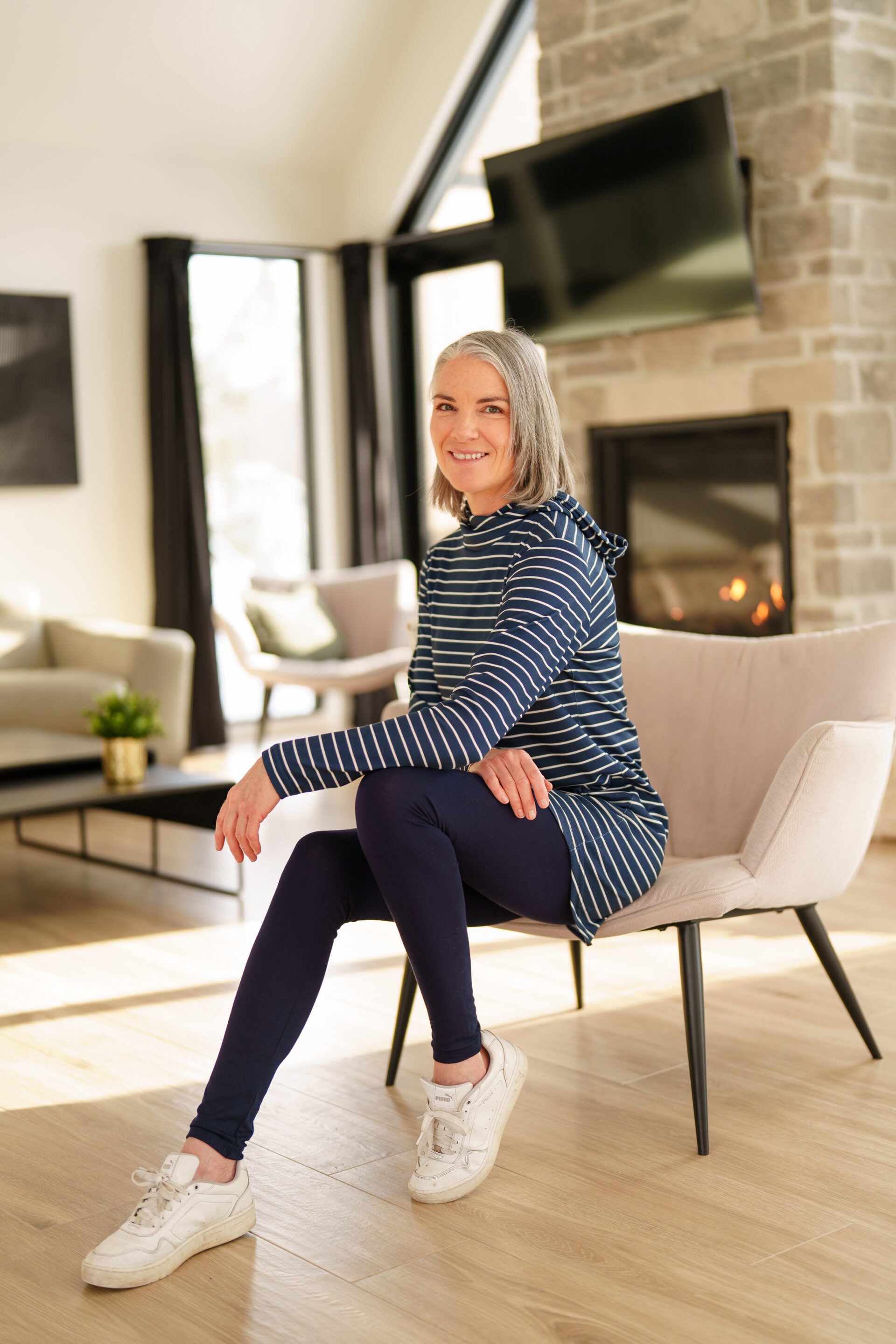 A woman wearing the Rupert Tunic by Rien ne se Perd in Navy/Ivory stripe, with  a crossover neckline and hood, long sleeves, and rounded hem. She is wearing it with black leggings and standing in a living room. 