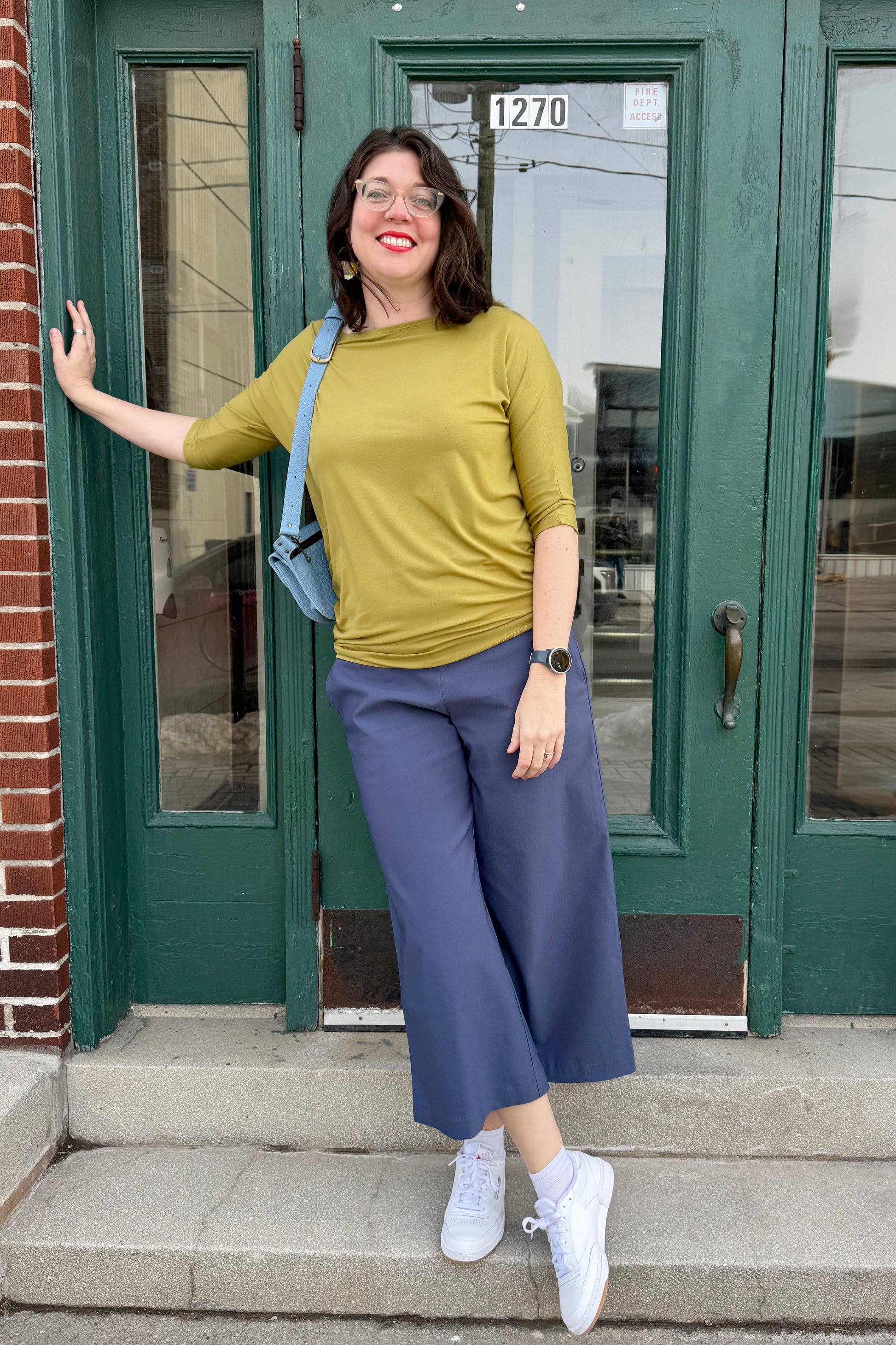Woman standing on steps in front of a green door, wearing the Bettina top by Moovment in Limette and blue pants.