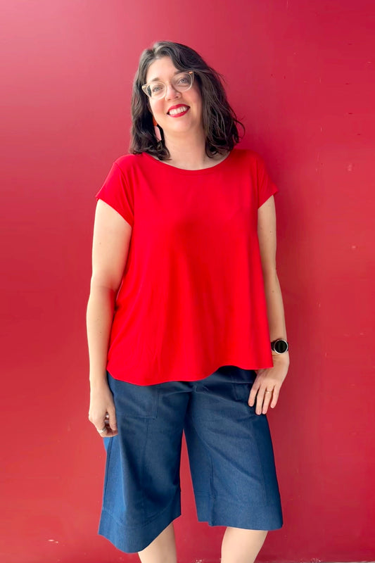 A woman wearing the Brooke Capri by Julei, wide-legged denim capris with a white drawstring and elastic at the back waist. She is wearing them with  a red shirt and standing against a red wall.