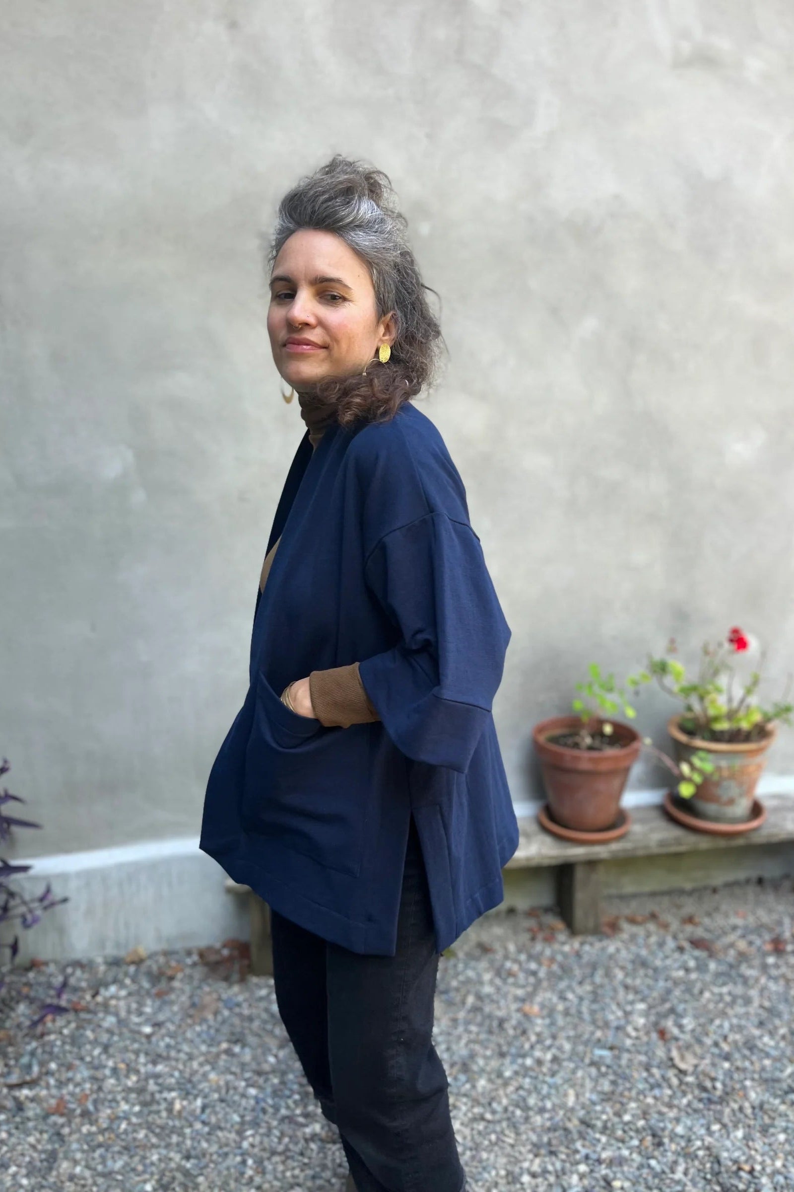 Side view of a woman wearing the Cydonia Cardigan by Kazak in Navy, a hip-length open cardigan with 3/4 kimono sleeves and large patch pockets. She is wearing it with black pants and a black top and standing outside.