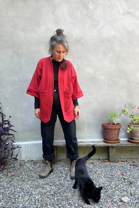 A woman wearing the Cydonia Cardigan by Kazak in Brick, a hip-length open cardigan with 3/4 kimono sleeves and large patch pockets. She is wearing it with black pants and a black top and standing outside with a black cat. 