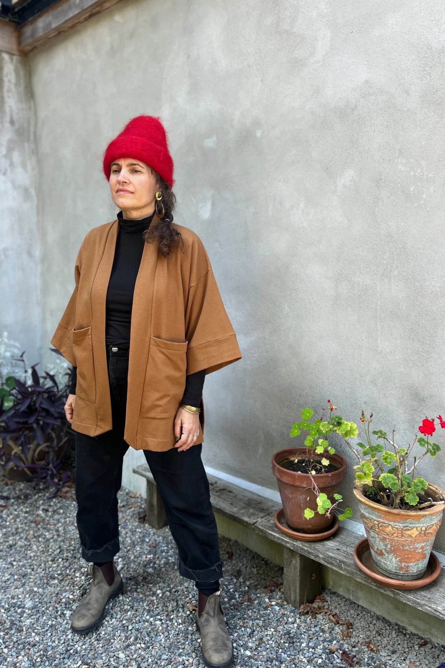 A woman wearing the Cydonia Cardigan by Kazak in Teak, a hip-length open cardigan with 3/4 kimono sleeves and large patch pockets. She is wearing it with black pants and a black top and standing outside.